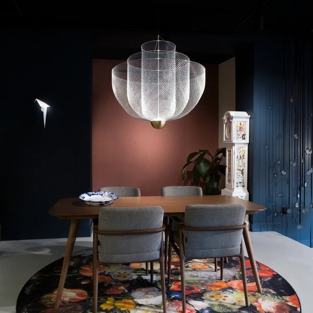 A Clérachic mesh chandelier lit in a dark blue room with a coral panel, above a wooden table with grey chairs and a flower-print rug.