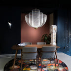 A Clérachic mesh chandelier lit in a dark blue room with a coral panel, above a wooden table with grey chairs and a flower-print rug.