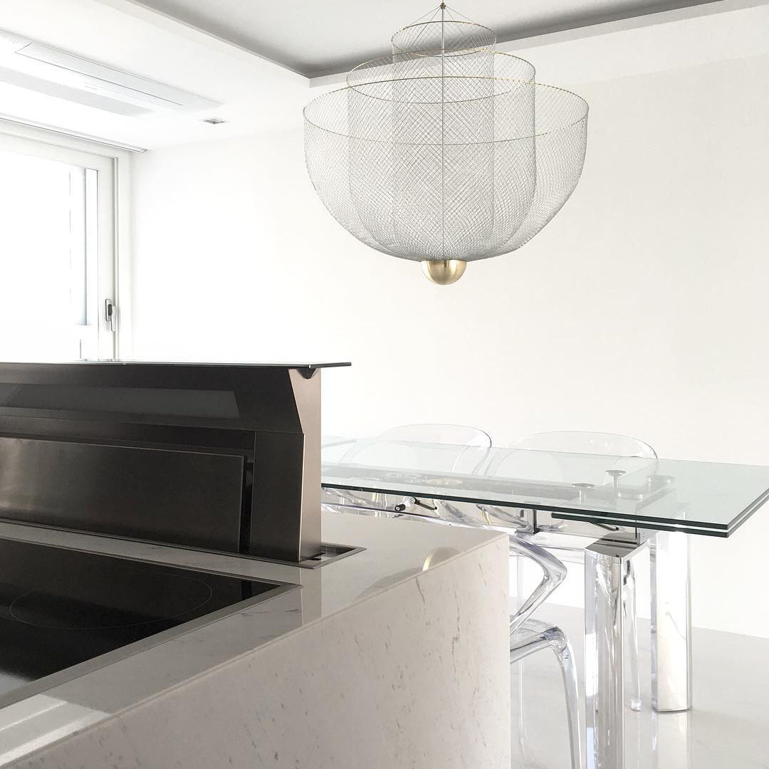 Clérachic wire-mesh chandelier in a white room with a glass table and a tall, ghostly chair, partly seen behind a counter.