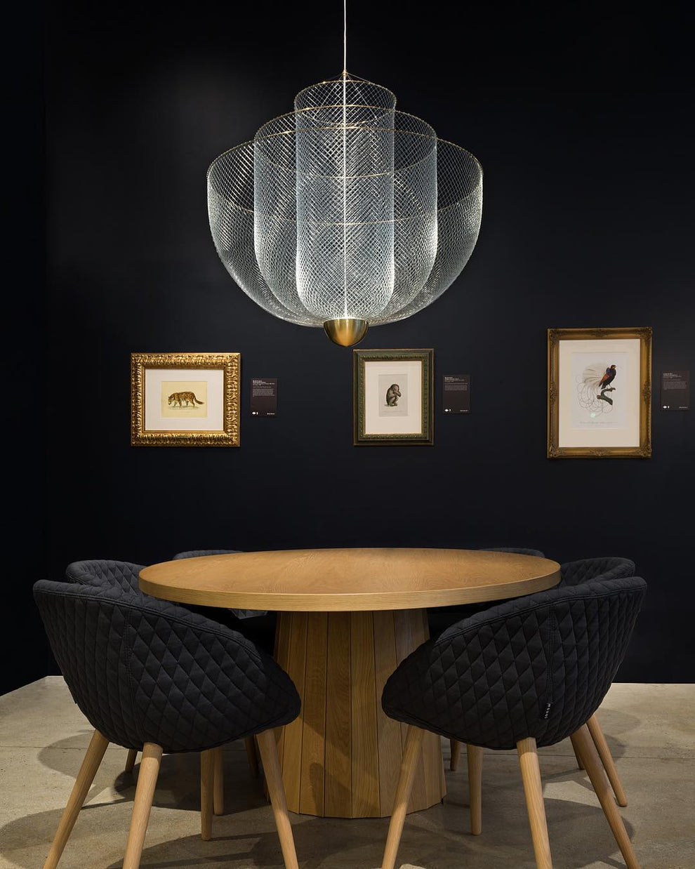A Clérachic mesh chandelier lit above a round wooden table and black quilted chairs, in front of a navy wall with framed pictures.