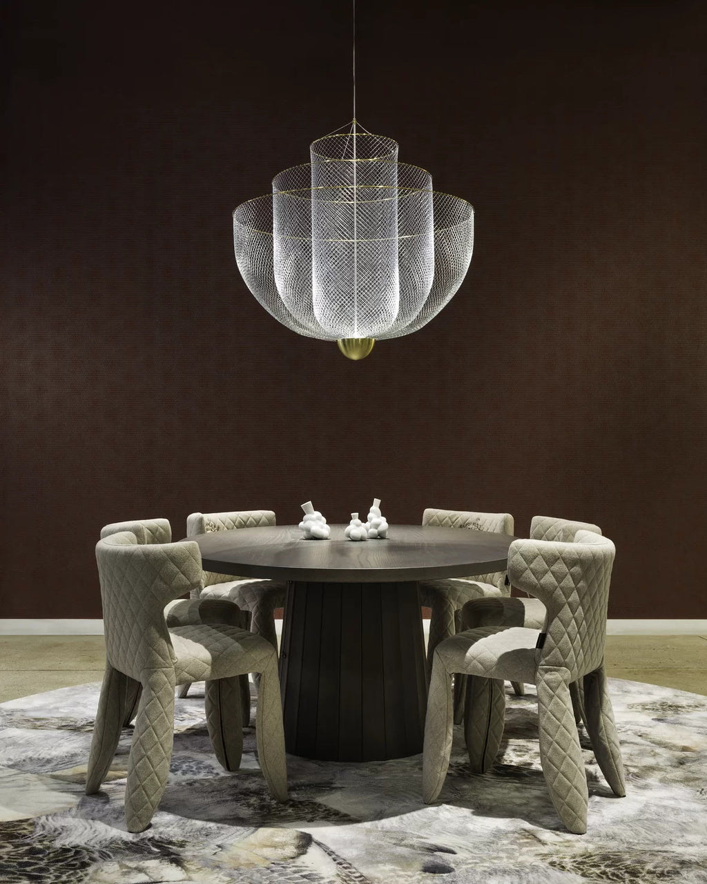 Clérachic silver wire-mesh chandelier above a dark round table with six upholstered chairs, on a patterned rug against a dark brown wall.