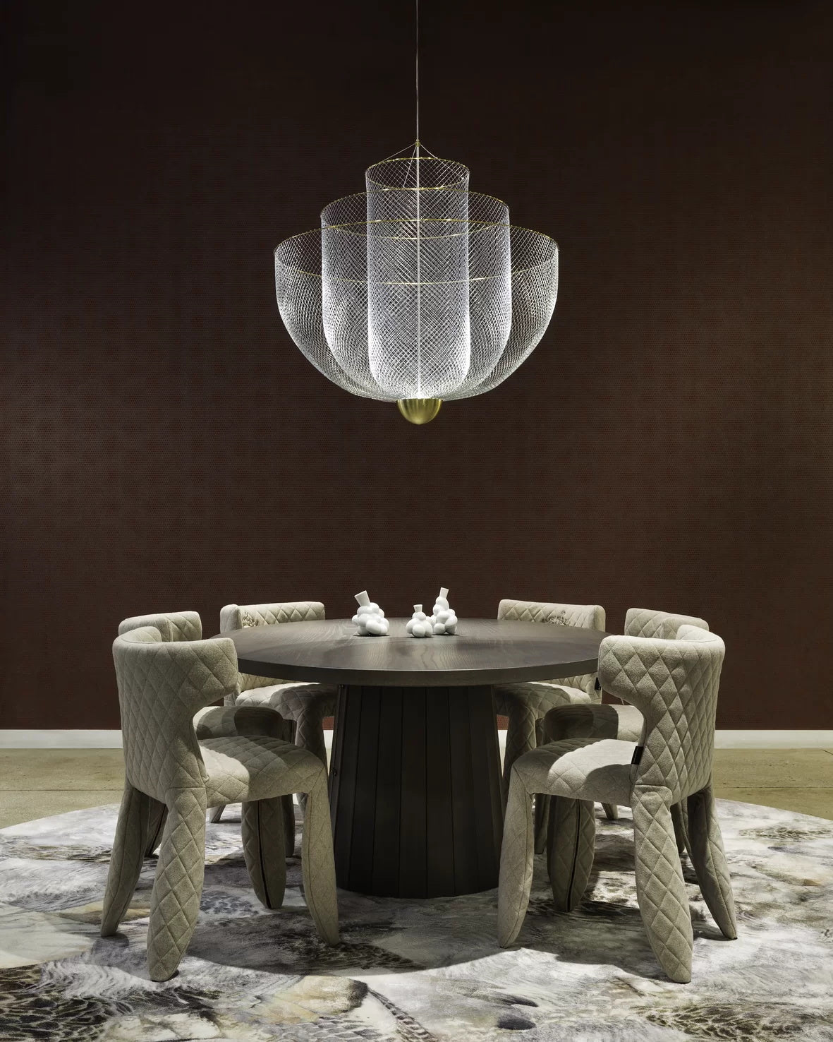 Clérachic silver wire-mesh chandelier above a dark round table with six upholstered chairs, on a patterned rug against a dark brown wall.