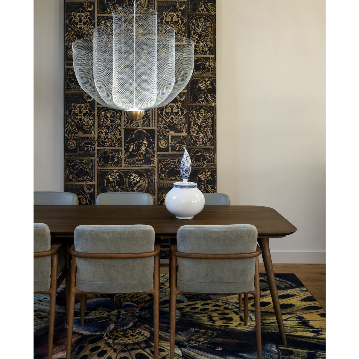 A Clérachic mesh chandelier over a walnut dining table with grey chairs, in front of a dark patterned wall hanging and a blue vase.