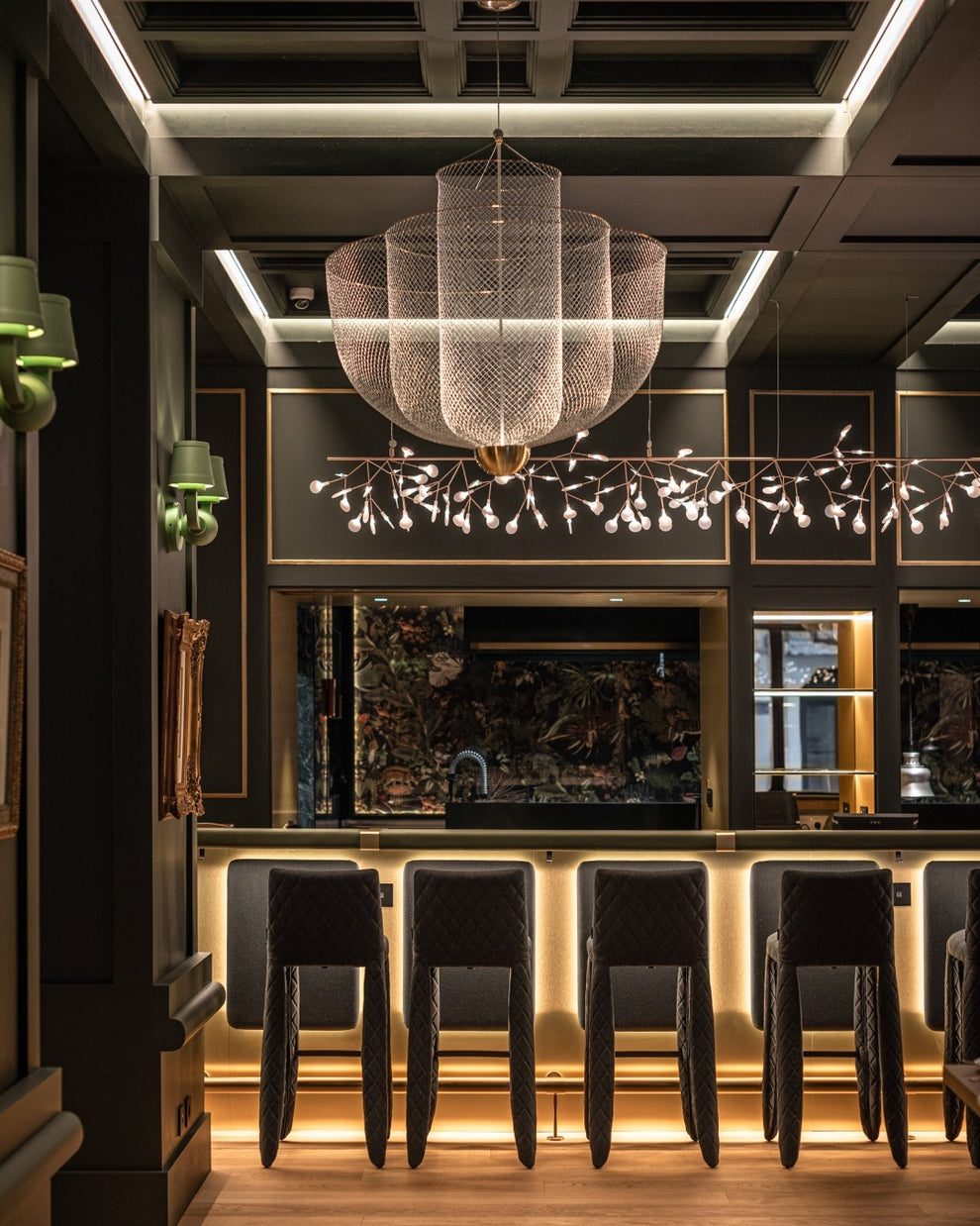 A Clérachic mesh chandelier in a dark bar, with a lit ceiling grid and a row of black stools below.