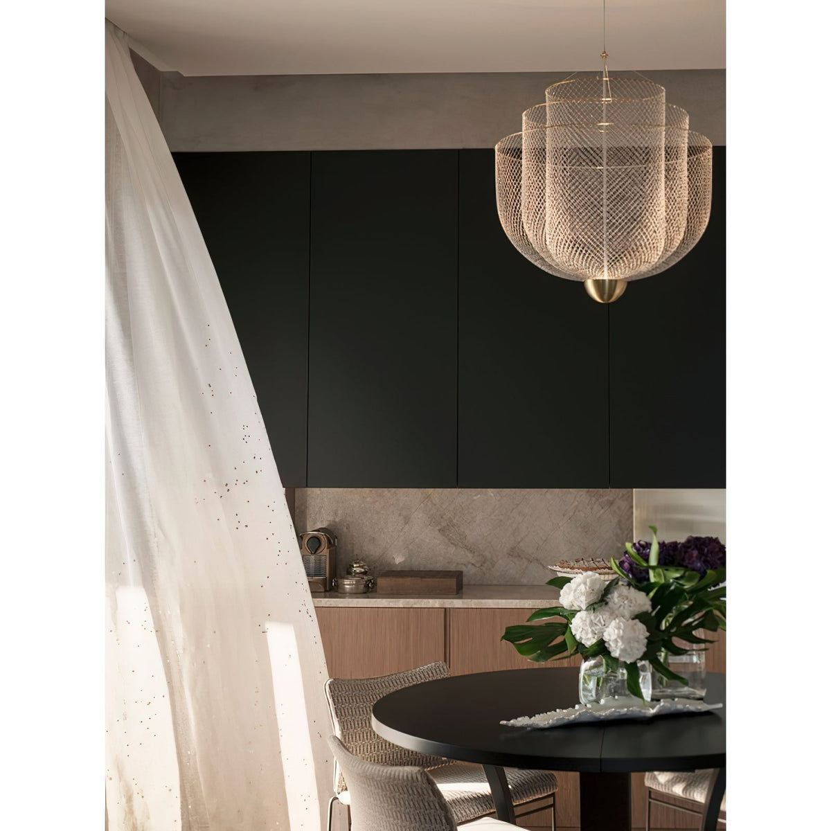 A Clérachic mesh chandelier in a kitchen and dining space with dark cabinets, a white curtain and a bouquet of hydrangeas.