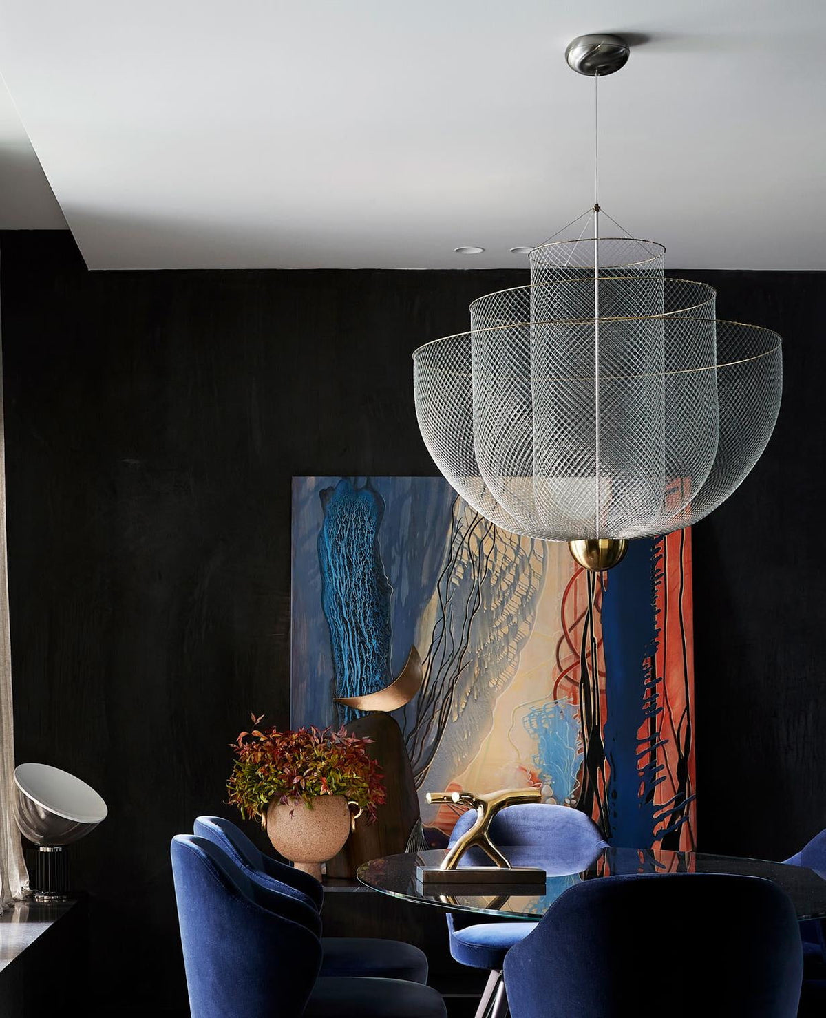 Clérachic wire-mesh chandelier in a dark room with a large abstract painting, blue velvet chairs and a small plant.