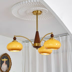 Clérachic Wood and Brass Chandelier in amber, three shades lit beside sheer curtains and a gilt frame.