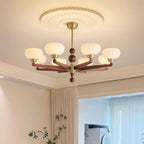 A Clérachic chandelier lit beneath a ribbed ceiling rose, with a corner of the room and a leafy branch in the background.