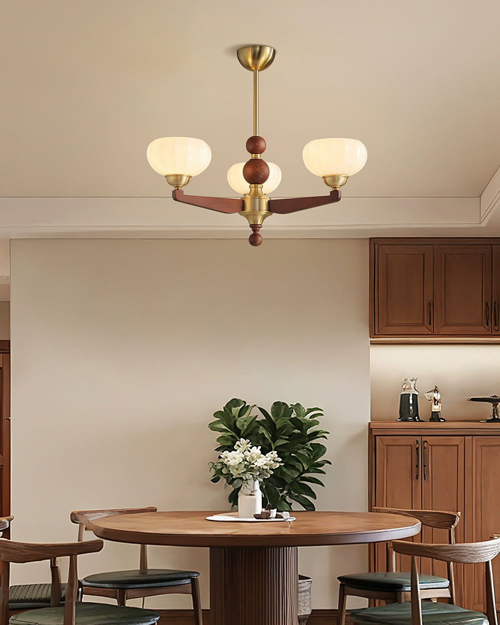 A smaller three-light Clérachic chandelier hanging above a round wooden dining table with a plant, dark chairs and wooden cabinets beyond.