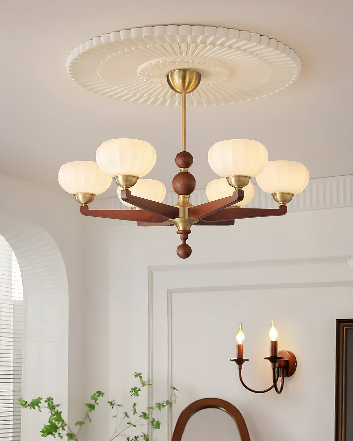 This Clérachic chandelier hangs close to a ribbed ceiling rose, with a candle-style wall light and a leafy plant in the room below.