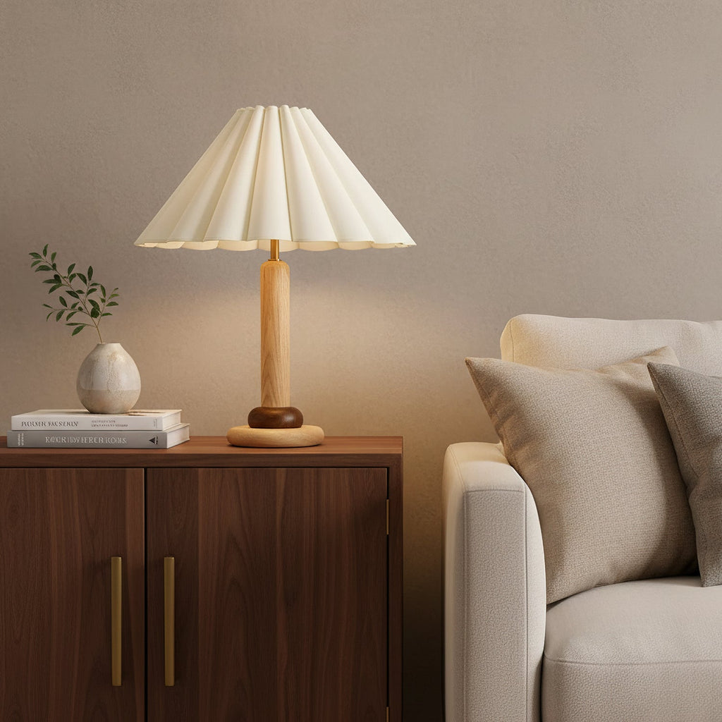 A Clérachic pleated table lamp sits on a walnut-toned cabinet beside a cream sofa and a small plant in a pale vase.