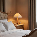 Lit warmly on a carved bedside table, this Clérachic pleated lamp sits beside a tufted headboard, white bedding and heavy curtains.