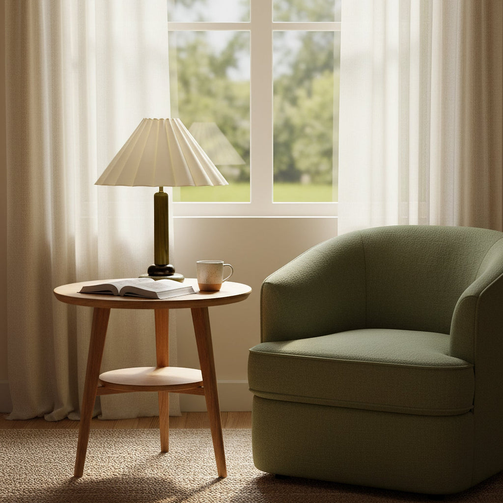 A Clérachic table lamp with a dark olive stem stands on a round side table by a window, next to a green armchair and an open book.