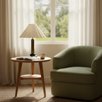 A Clérachic table lamp with a dark olive stem stands on a round side table by a window, next to a green armchair and an open book.