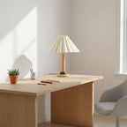 A Clérachic pleated table lamp sits on a light wood desk in a bright room, beside a small plant, a mug of pens and a notebook.