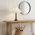 An olive-green-stemmed Clérachic table lamp stands on a pale wood console under a round black-framed mirror, with a basket of dried cotton stems below.