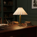 A lit Clérachic table lamp with a dark green stem sits on a dark wood desk by shelves of books, with glasses and a quill pot.