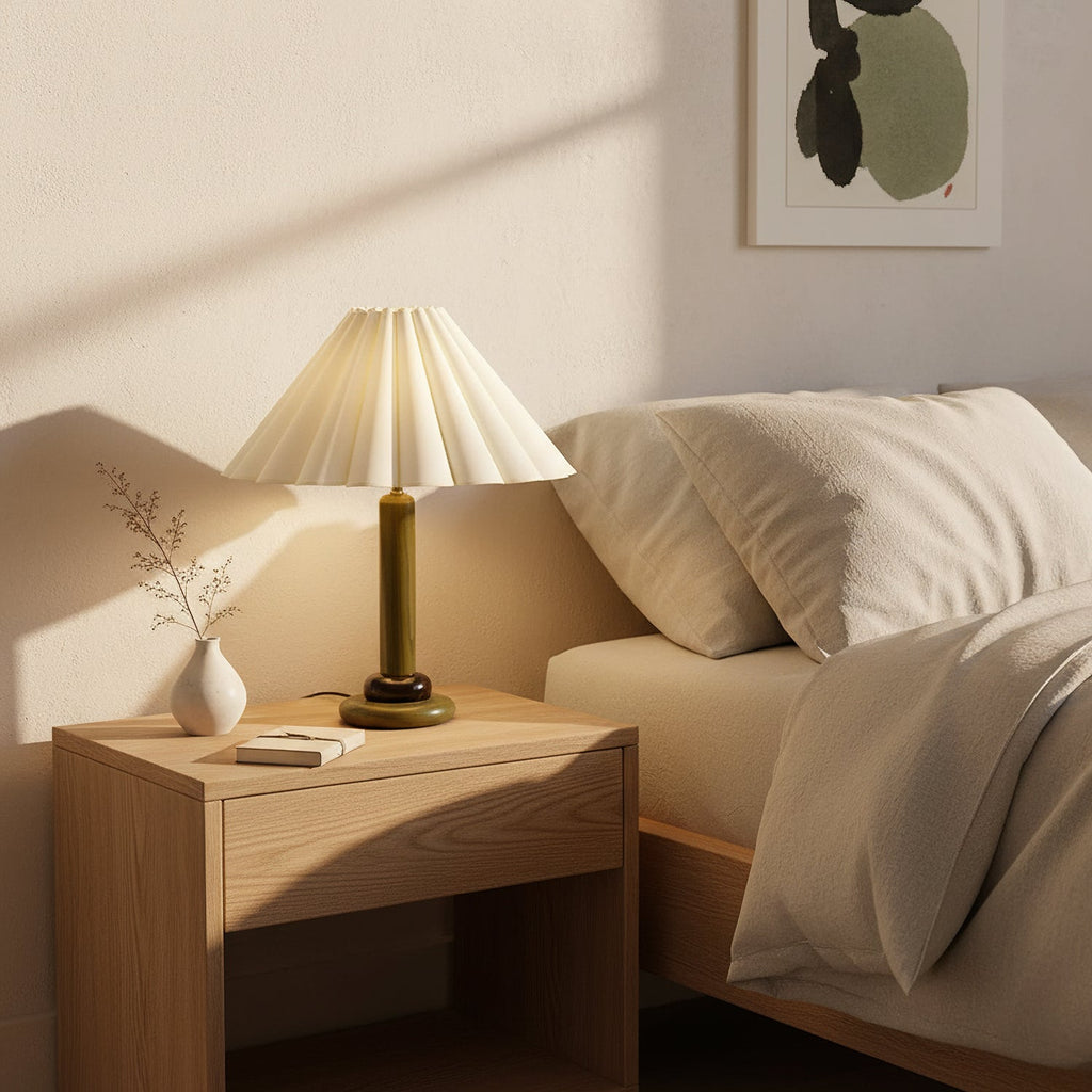On a light wood bedside table, a lit Clérachic table lamp with an olive-green base stands beside a bud vase and a made bed.
