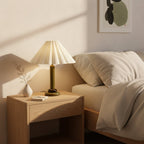 On a light wood bedside table, a lit Clérachic table lamp with an olive-green base stands beside a bud vase and a made bed.