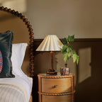 A Clérachic pleated lamp with a dark wood stem stands on a round carved bedside table, beside a bobbin-turned headboard and a small ivy plant.