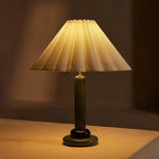 Glowing amber in a dim room, this Clérachic table lamp has a green stem, a dark ring and a pleated shade lit from inside.