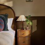 A Clérachic table lamp with a dark wood stem sits low on a carved round bedside table next to a turned wooden headboard and an ivy plant.