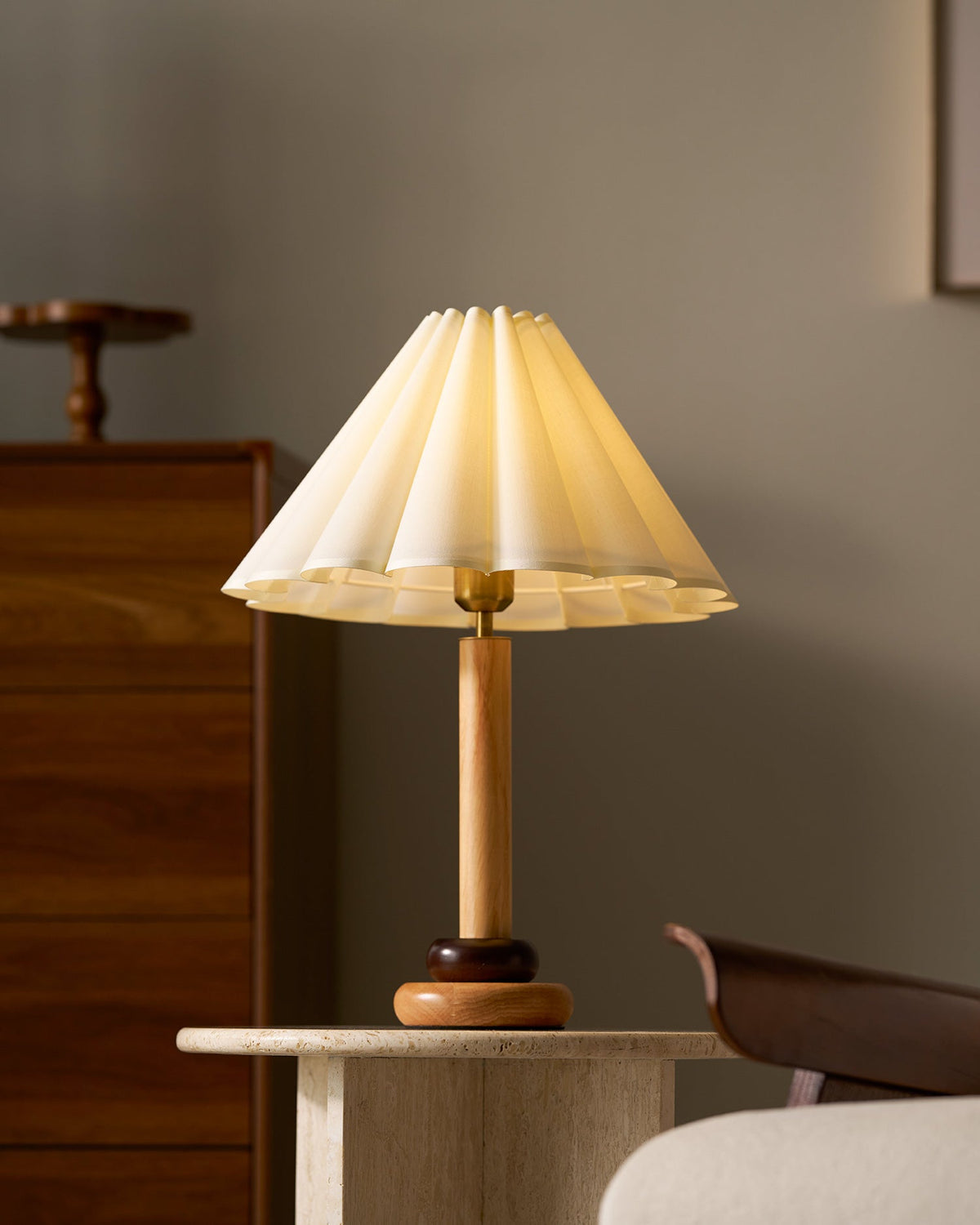 Lit and glowing, this Clérachic pleated table lamp sits on a travertine side table beside a walnut chest of drawers and a cream armchair.