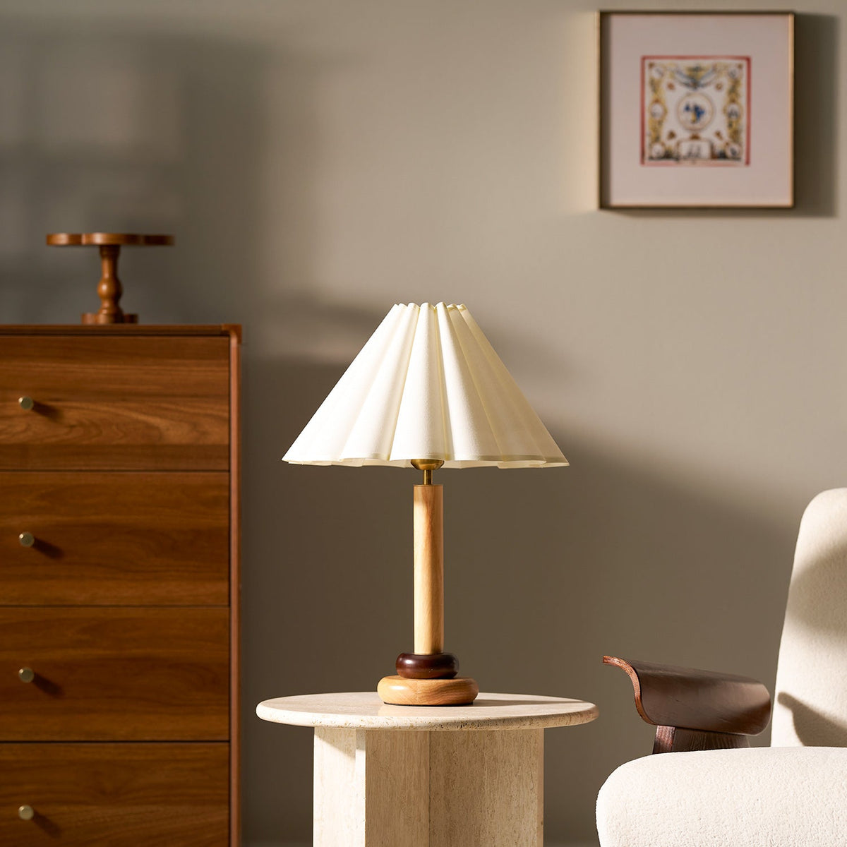 Seen from slightly below, this Clérachic pleated table lamp stands on a round stone table beside a walnut chest and a cream chair.