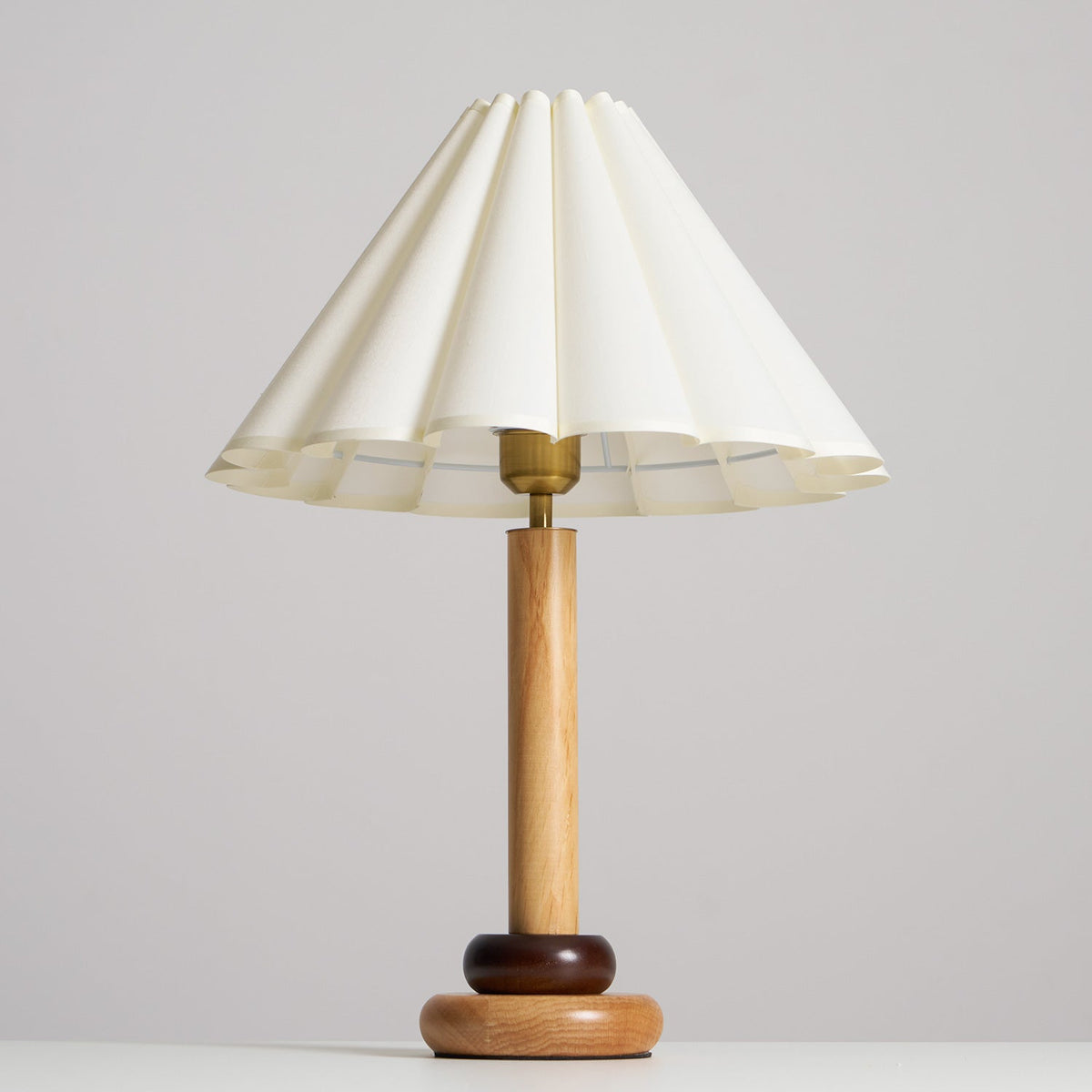 A Clérachic table lamp with a natural wood stem, a brass fitting and a wide scalloped cream shade against a pale grey backdrop.