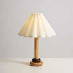Lit softly, this Clérachic table lamp has a pleated cream shade, a slim wood stem and a stepped base on a pale tabletop.