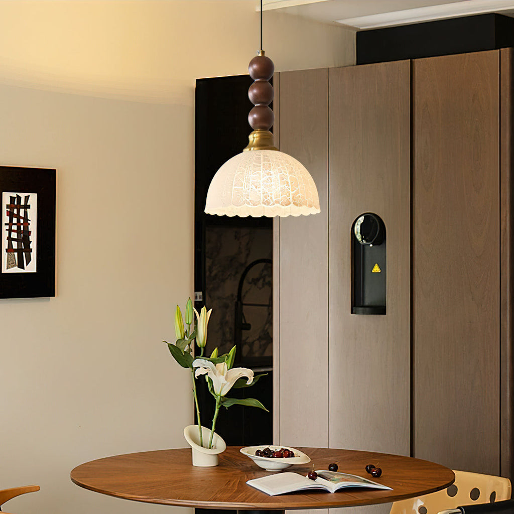 A Clérachic pendant with a lit, scalloped frosted shade and walnut beads hangs over a dining table set with flowers.
