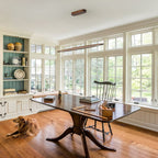 A slim Clérachic wood pendant hangs in a sunroom with tall windows, above a dark wooden table with a spindle chair.