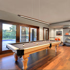 A Clérachic wood pendant hangs over a billiards table in a room with wooden floors and tall glass doors.