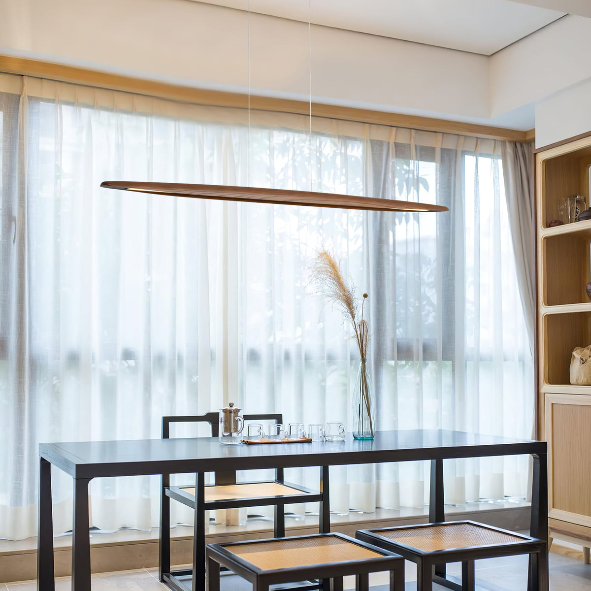 A dark walnut Clérachic pendant hangs in front of sheer curtains above a black table, with a grass stem in a vase.