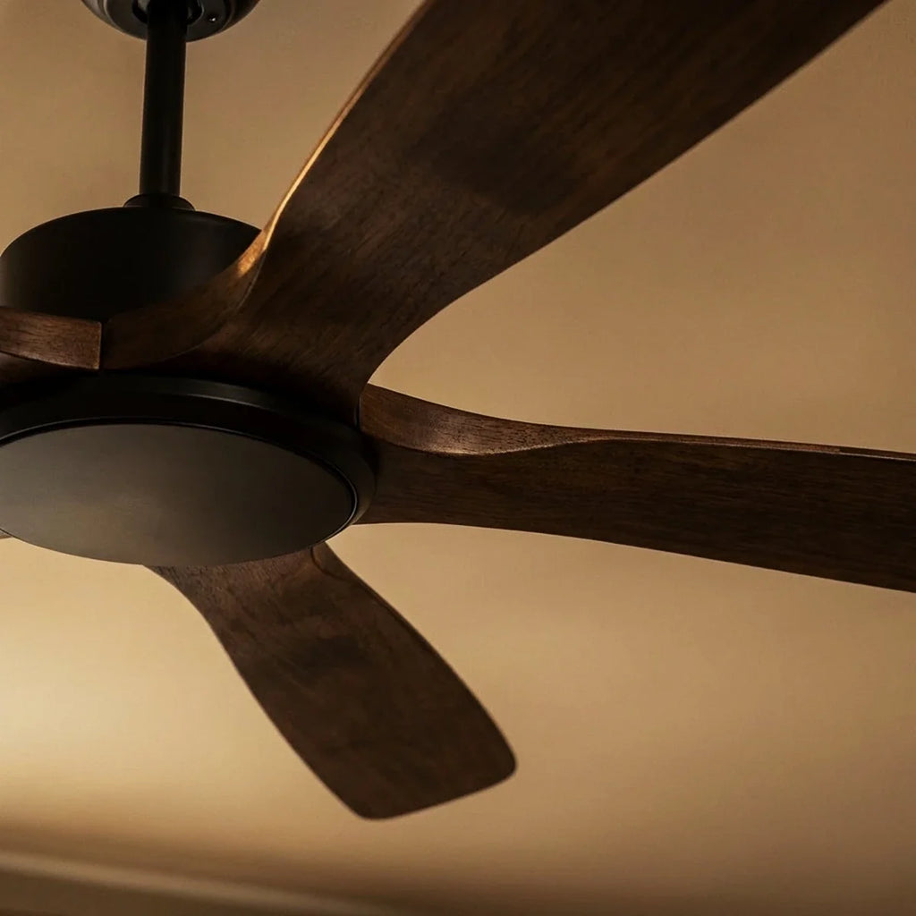 Low angle on the Clérachic fan's black motor, dark light disc and rich walnut-toned blades glowing under warm ceiling light.