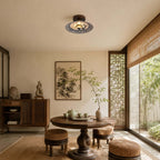 A Clérachic ceiling light above a round wooden table and woven stools in a bright room with a lattice screen and garden view.