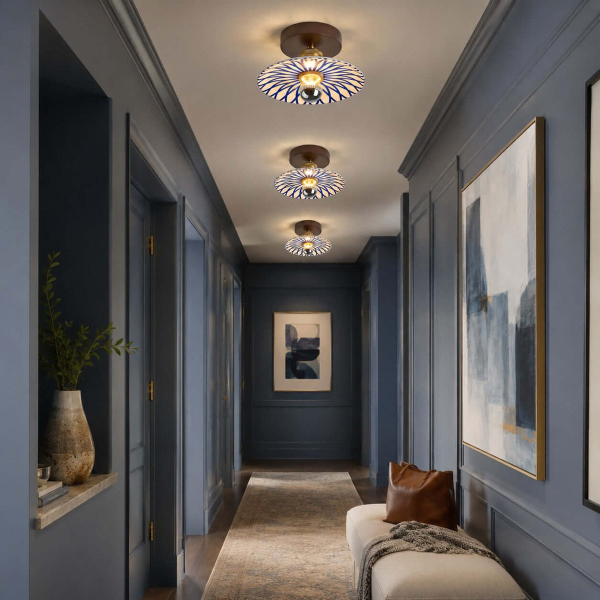 Three Clérachic ceiling lights lead down a dark blue hallway, with a large abstract painting and a cream bench.