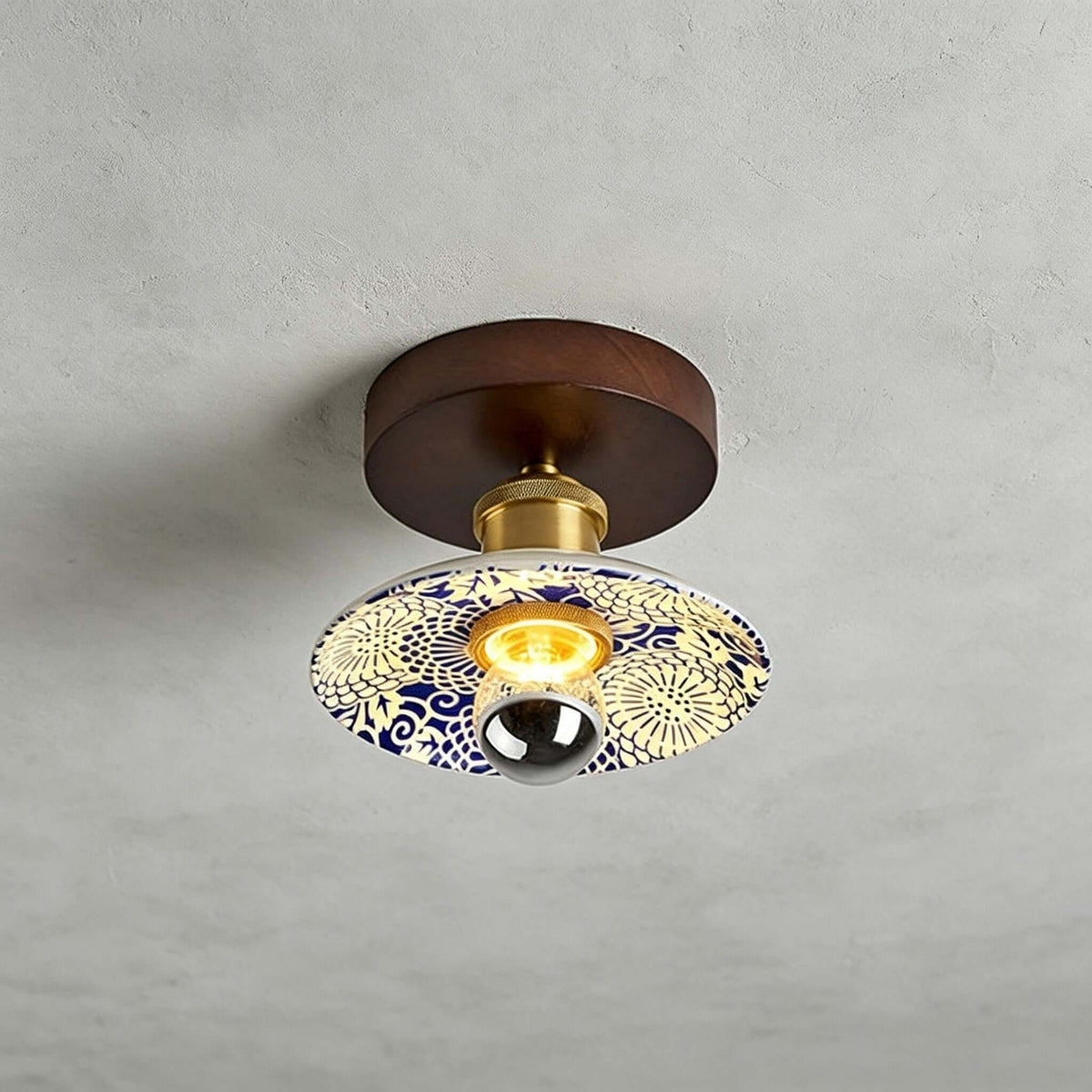 A Clérachic ceiling light with a cream shade patterned in blue and yellow, a round wood canopy and a brass fitting above.