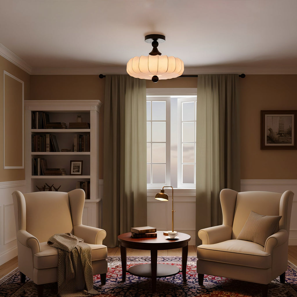 A lit Clérachic ceiling light with a beige pleated shade hangs in a warm study with two wing chairs, olive curtains and a patterned rug.