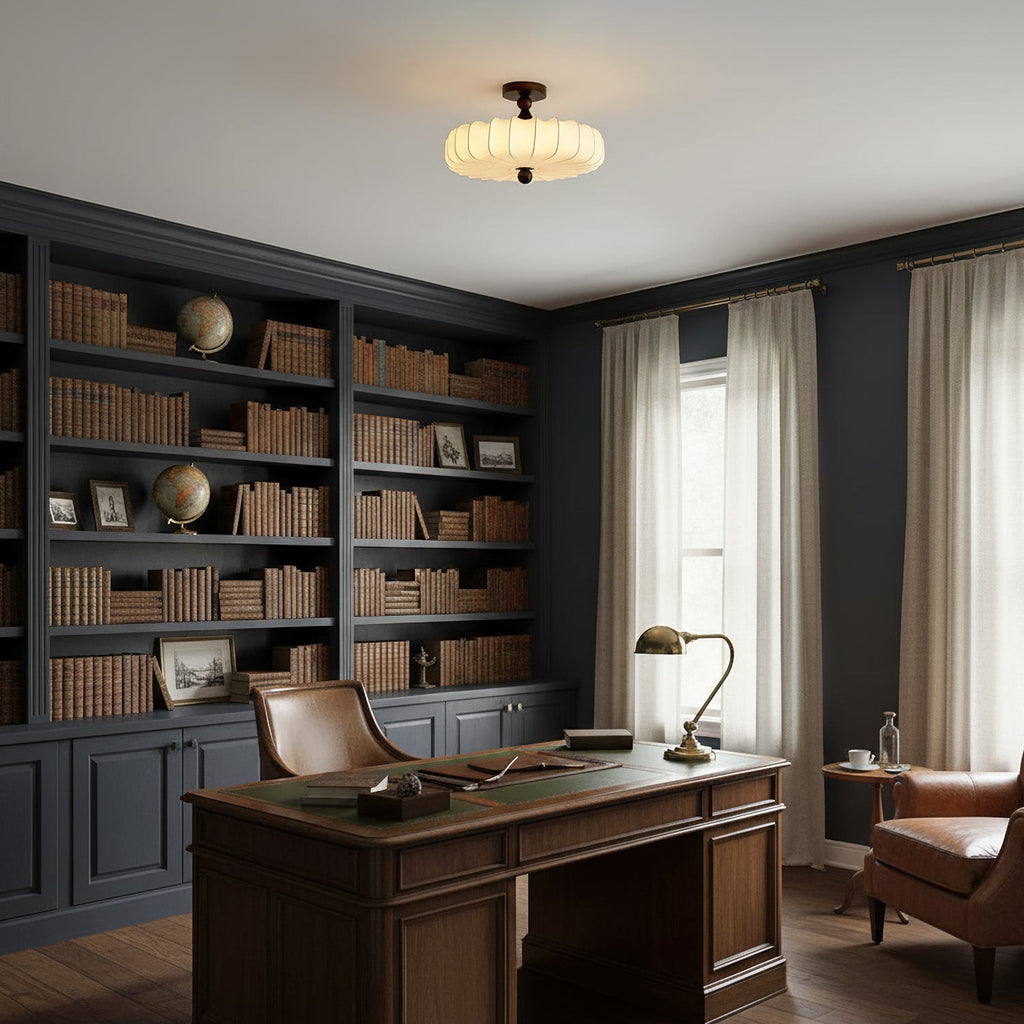A lit Clérachic ceiling light with a ribbed fabric shade hangs in a study with dark shelving, a leather chair and a wooden desk.