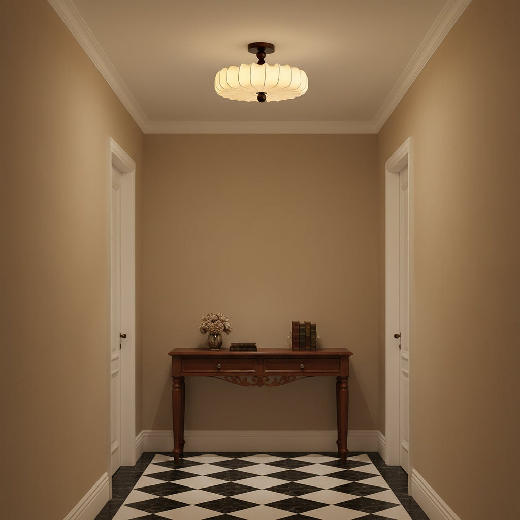 A glowing Clérachic ceiling light with a pleated shade lights a small tan-walled hallway with a carved console table and a chequered floor.