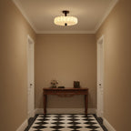 A glowing Clérachic ceiling light with a pleated shade lights a small tan-walled hallway with a carved console table and a chequered floor.