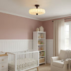 In a pink nursery with a white cot and shelving, a lit Clérachic ceiling light with a soft pleated shade hangs from the ceiling.