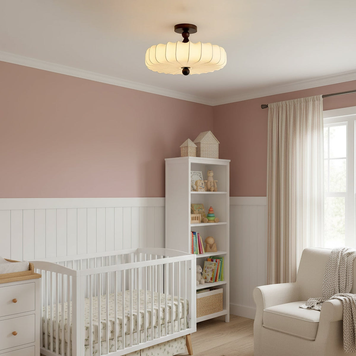 In a pink nursery with a white cot and shelving, a lit Clérachic ceiling light with a soft pleated shade hangs from the ceiling.