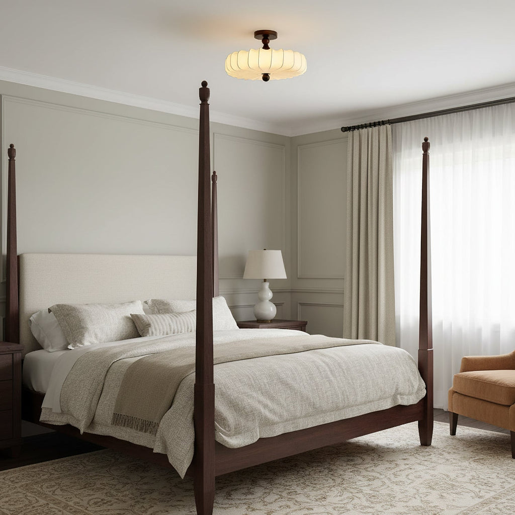 A small Clérachic ceiling light with a pale pleated shade hangs high above a four-poster bed in a calm grey-beige bedroom.