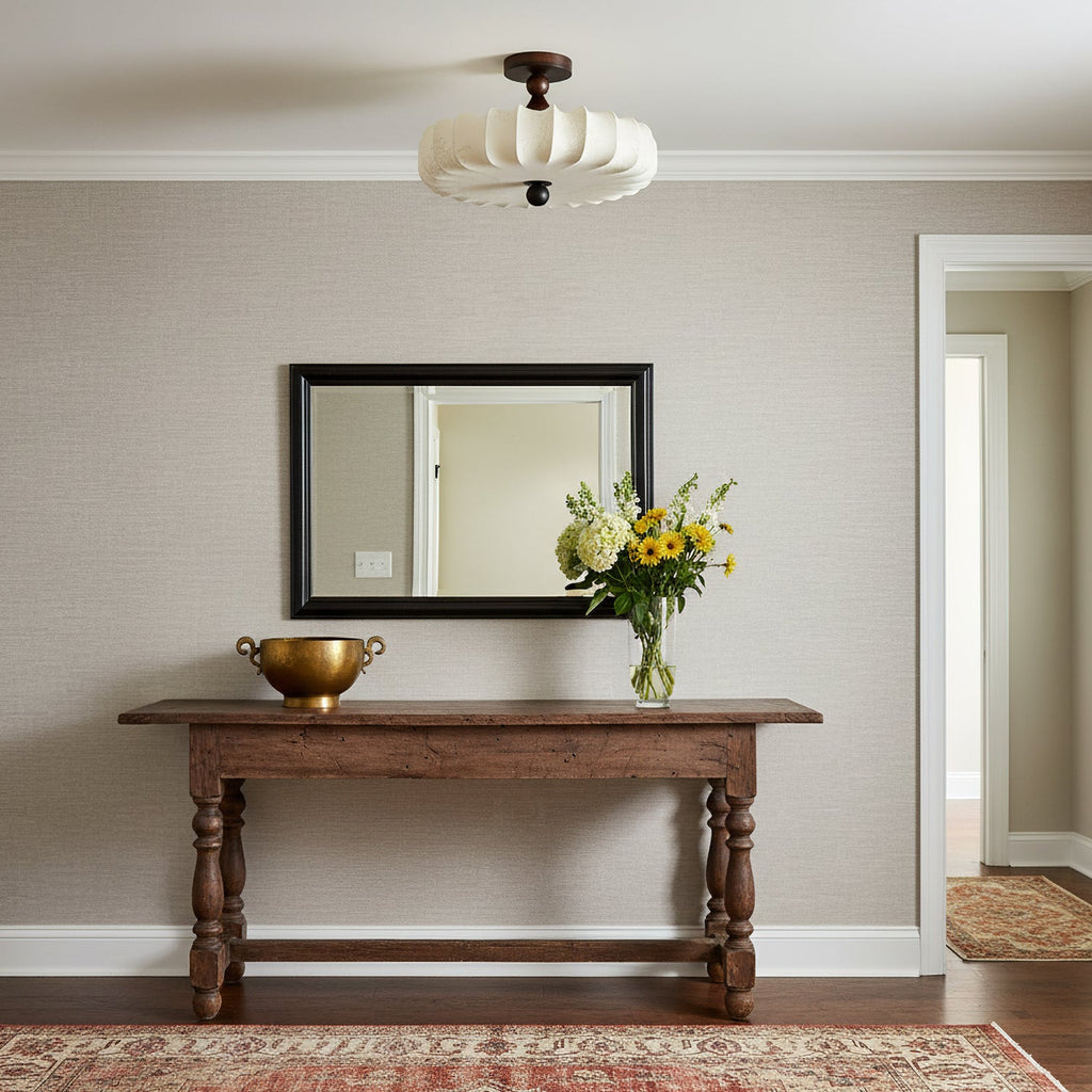 An unlit Clérachic ceiling light with a cream pleated shade hangs above a carved wooden console table, a mirror and a bunch of yellow flowers.