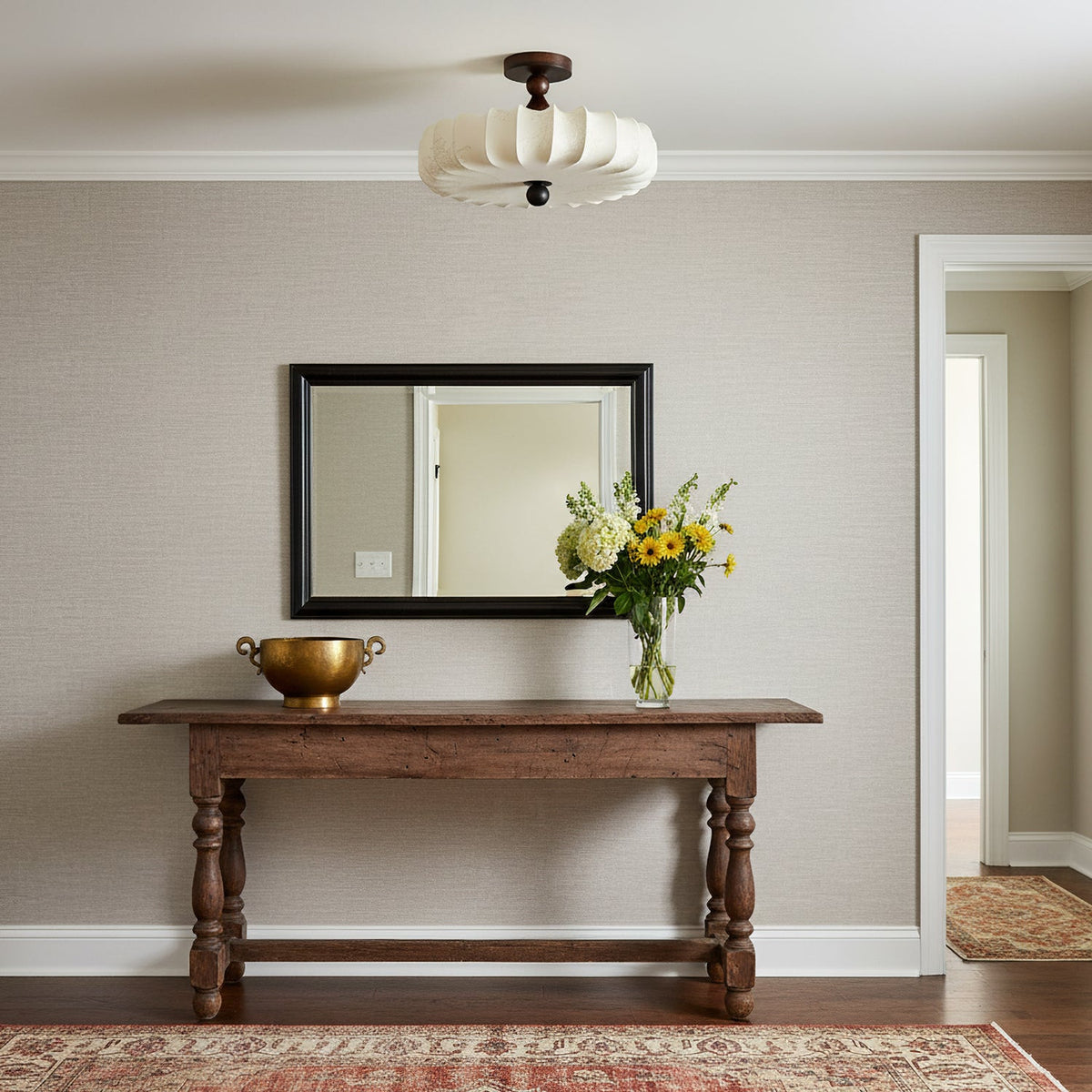 An unlit Clérachic ceiling light with a cream pleated shade hangs above a carved wooden console table, a mirror and a bunch of yellow flowers.