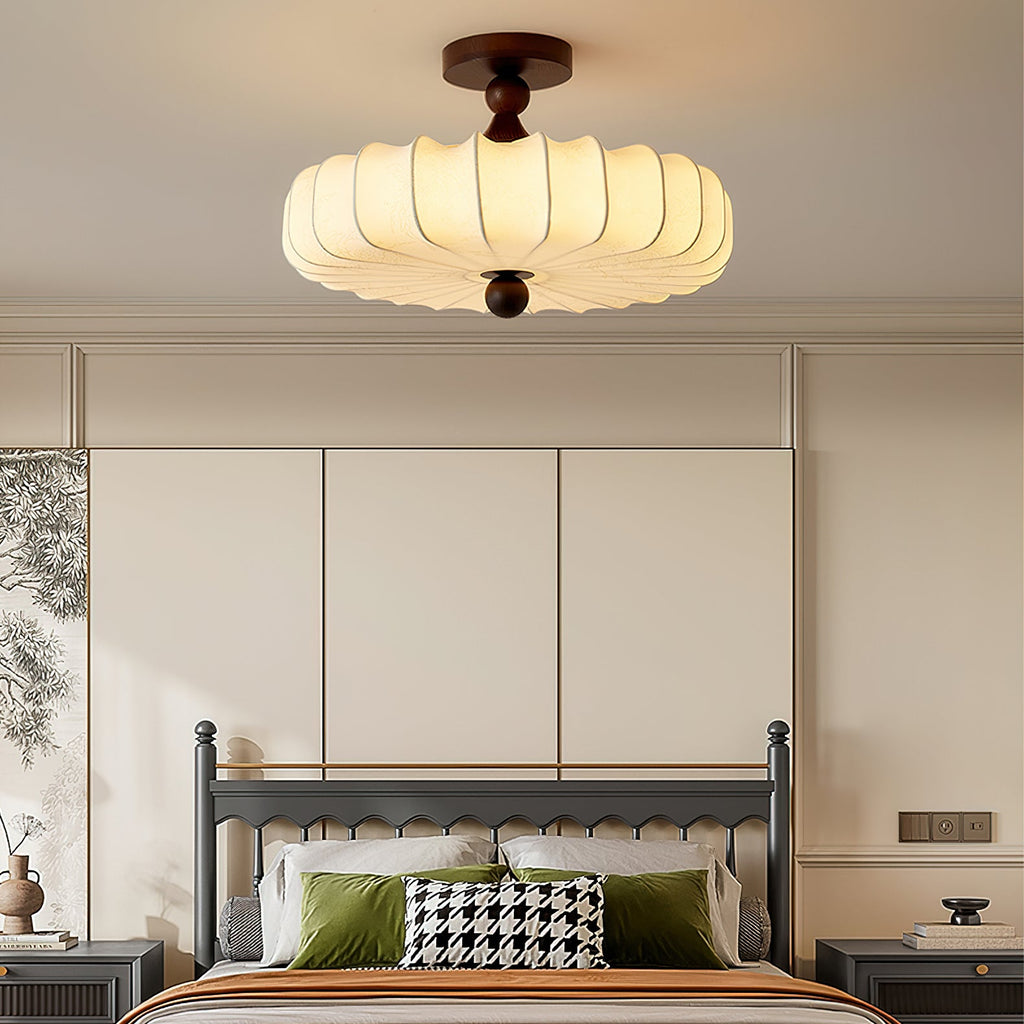 A large Clérachic ceiling light with a pleated cream shade glows above a bed with a grey frame, green cushions and a houndstooth pillow.