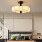 A large Clérachic ceiling light with a pleated cream shade glows above a bed with a grey frame, green cushions and a houndstooth pillow.
