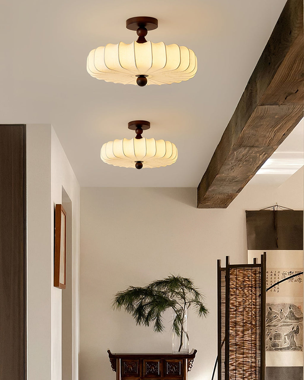 Two lit Clérachic ceiling lights with pleated shades hang along a ceiling with a dark wooden beam, above a small plant and a carved side table.